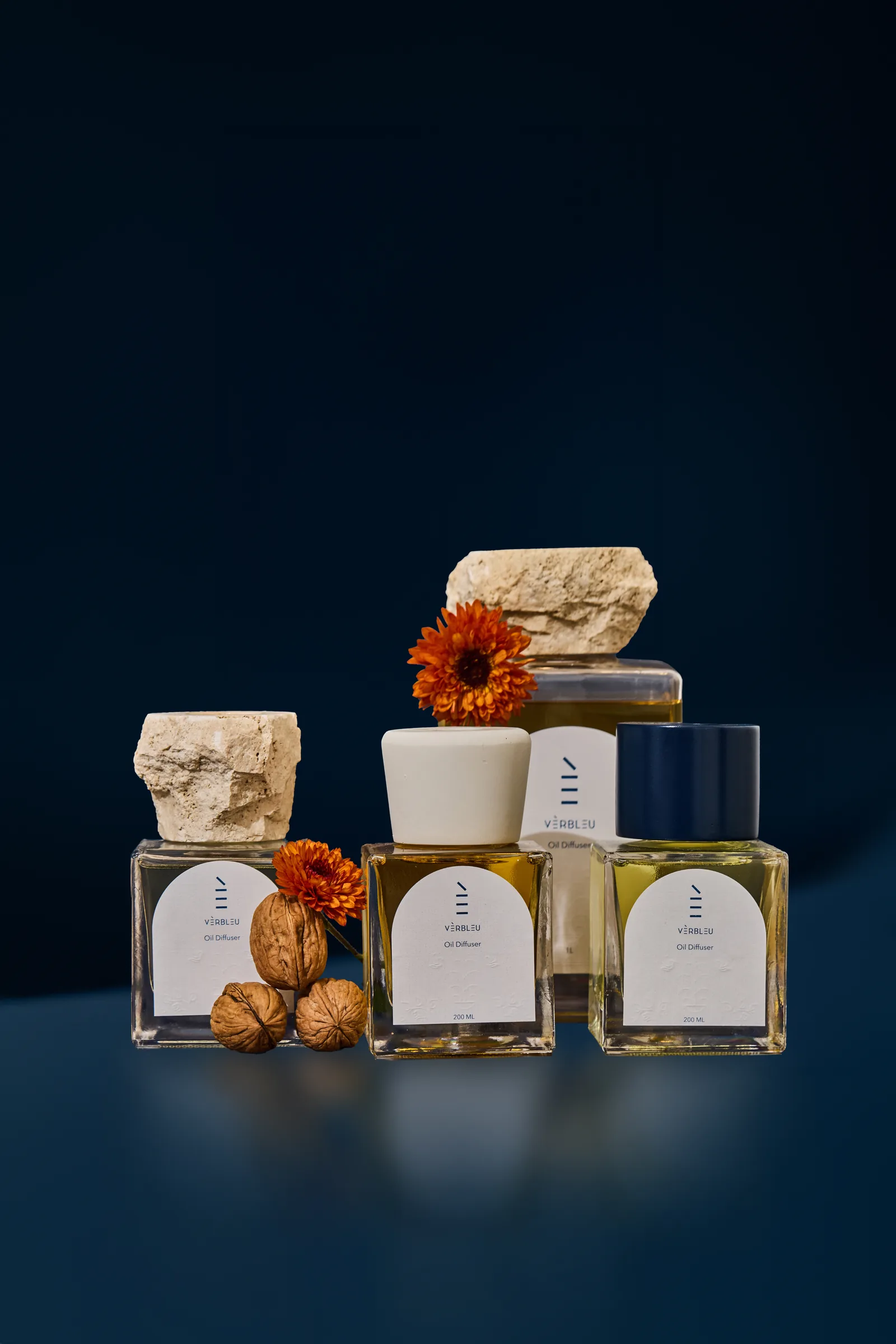 Verbleu oil diffusers with stone and ceramic lids, orange flowers and walnuts against a navy background